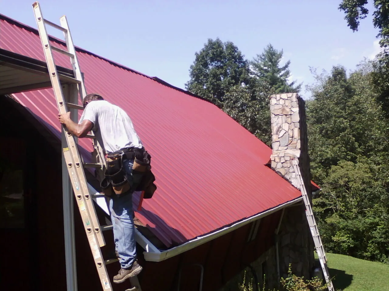 Roofer on a ladder beside a red metal roof and stone chimney — Soffit & Fascia Repair by Roofing Brothers in Millis